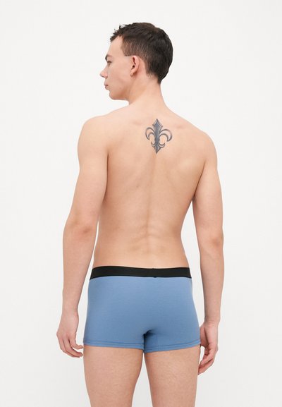 Boxeri albaștri dintr-un material texturat, cu un tiv elastic negru, punând în evidență o tatuaj în formă de floare-de-lis pe spate.