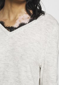 Jasnoszary sweter z dekoltem w kształcie litery V oraz czarną koronkową lamówką wzdłuż kołnierza. Miękka, fakturowana tkanina o luźnym kroju.