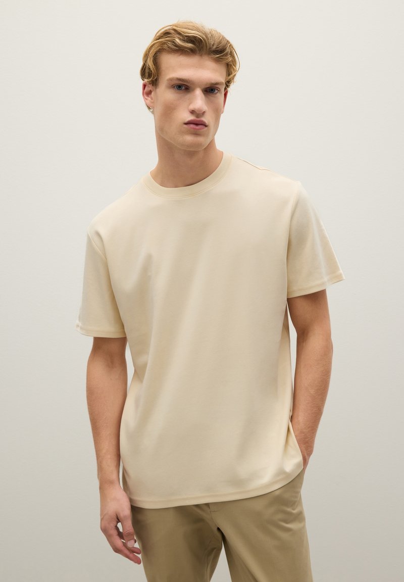 T-shirt beige a maniche corte in tessuto morbido con scollatura rotonda e vestibilità rilassata, indossato con pantaloni chiari, caratterizzato da un design minimale e senza fantasie.