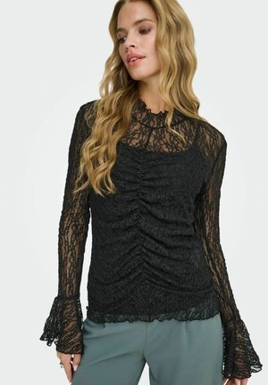 Kobieta o długich blond włosach, ubrana w prześwitującą czarną koronkową bluzkę z długim rękawem, marszczonym przodem i rozszerzanymi mankietami, w połączeniu z zielonymi spodniami.
