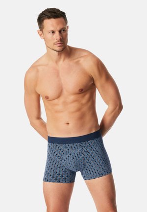 Mężczyzna z krótkimi włosami bez koszulki, ubrany w niebieskie bokserki w wzory, stojący z rękami za plecami na jednolitym tle.