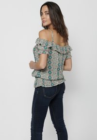 Blusa floreale off-shoulder con volant in beige chiaro, con motivi teal. Abbinata a jeans attillati in denim scuro.
