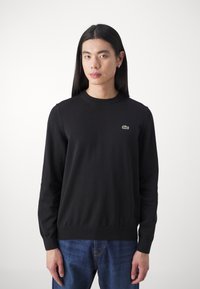 Sort striktrøje med rund halsudskæring og lange ærmer. Har et lille grønt Lacoste-logo på brystet. Bæres med blå jeans.