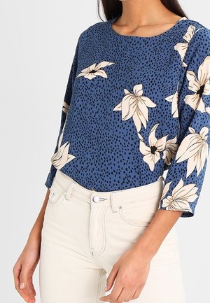 Femme portant un haut bleu à manches longues avec des motifs floraux crème et un pantalon beige clair, vue des épaules aux hanches sur un fond uni.