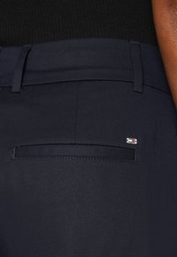 Pantaloni negri închisi, cu un buzunar la spate cu un mic logo de marcă. Material neted cu o croială ajustată și detalii cusute cu atenție.