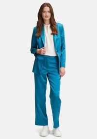 Turkos satin dräkt med en skräddarsydd kavaj och vida byxor, kombinerad med en vit blus med detaljer i nyckelhålet. Vita sneakers.