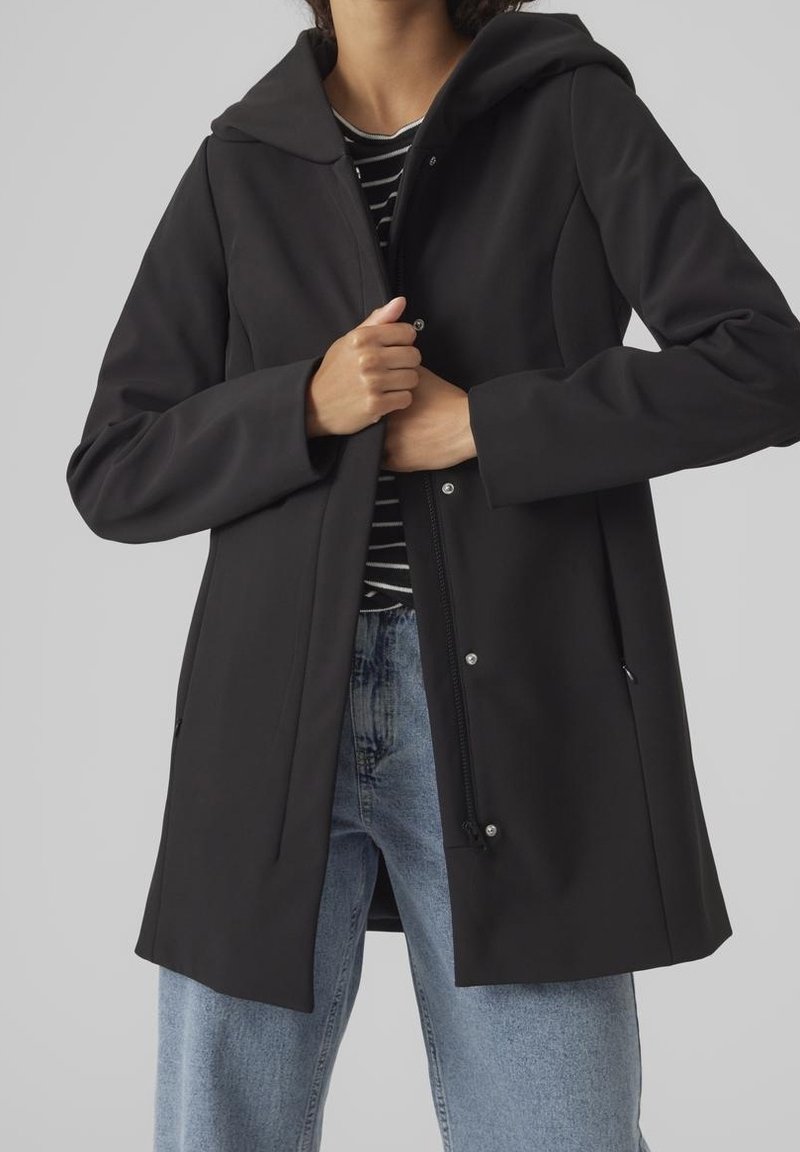 Chaqueta negra con capucha hecha de tela lisa, con botones a presión, cremallera frontal y bolsillos laterales, combinada con vaqueros azules casuales.