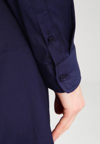 Marinblå långärmad skjorta med knäppta manschetter och mjuk bomullstextur, synlig sömnad och två mörka knappar.