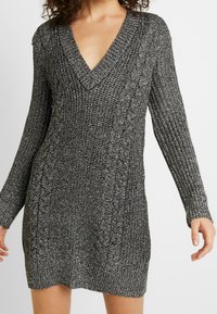 Robe à manches longues en tissu tricoté texturé noir et argent. Présente un décolleté en V profond et des détails en tricot torsadé sur les côtés.