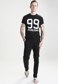 Must have mustat puuvillased T-särk, millel on valge tekst "99 PROBLEMS," koos mustade pükstega. Mees kannab mustrilisi sandaale. Nahalt on nähtavad tätoveeringud.