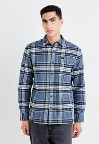 Camisa de franela de cuadros azul con cuello abotonado, mangas largas, adornos de patrón blanco y un bolsillo en el pecho con un pequeño logo.