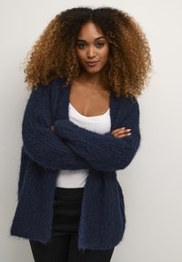 Tumši jūras zila kardigana ar pūkainu tekstūru, atvērtu priekšpusi un platām piedurknēm, kārtota virs baltas blūzes un apvienota ar melniem biksēm.