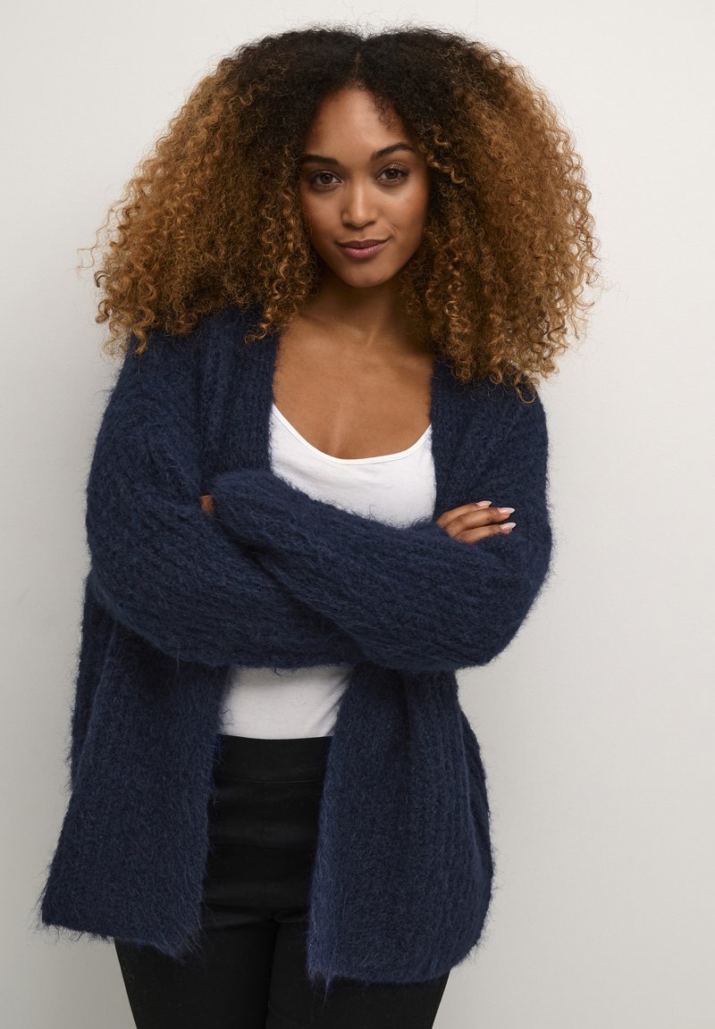 Tumši jūras zila kardigana ar pūkainu tekstūru, atvērtu priekšpusi un platām piedurknēm, kārtota virs baltas blūzes un apvienota ar melniem biksēm.