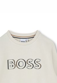 Sweatshirt beige clair avec un col ras du cou, présentant un grand logo "BOSS" en lettres noires et beige en contour. Texture en coton.