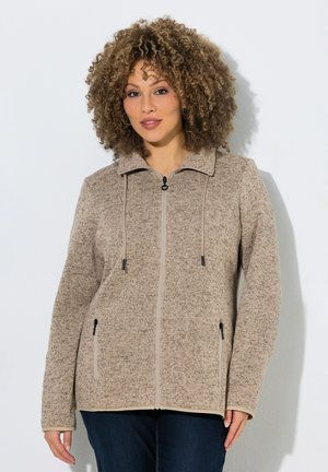 Kvinde med krøllet hår iført beige jakke med lynlås og sidelommer samt mørke jeans, stående foran en ensfarvet hvid baggrund.