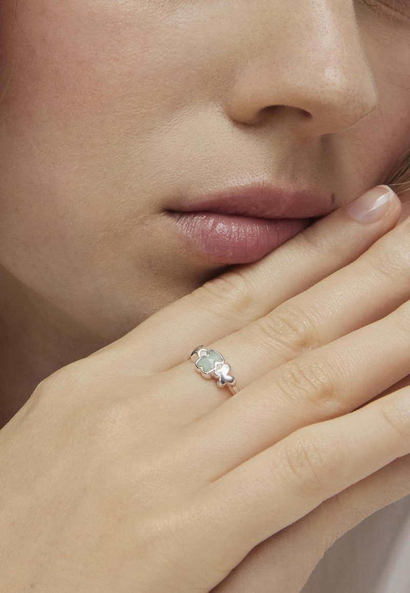 Anillo de plata con una piedra verde claro, diseñada con un patrón floral y textura suave, llevado en una mano cerca de un rostro.