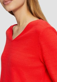 Top rosso con texture e scollatura a V. Il materiale presenta un motivo sottile, creando un effetto tridimensionale. Si adatta aderente al corpo.