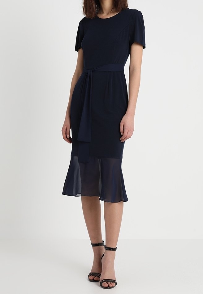 Vestido Marino con mangas cortas, una cintura con lazo y un dobladillo superpuesto de tul. Hecho de una tela suave; presenta una parte inferior fluida. Sandalias negras con tira al tobillo.