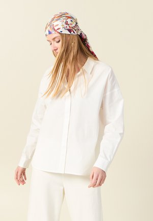 Chemise blanche à manches longues en tissu lisse. Une écharpe à motifs colorés est nouée autour de la tête. Fond clair.