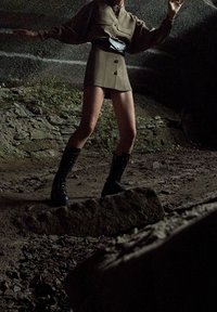 Persona che indossa un completo marrone con una borsa a vita e stivali neri, in piedi su un terreno roccioso in una grotta o un tunnel scarsamente illuminato.
