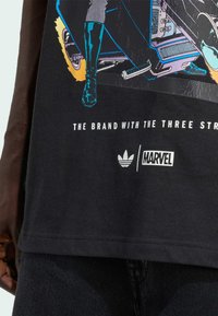 Nærbillede af sort Marvel-tema T-shirt med en farverig motorcykelgrafik, Adidas- og Marvel-logoer, samt delvis slogan tekst.