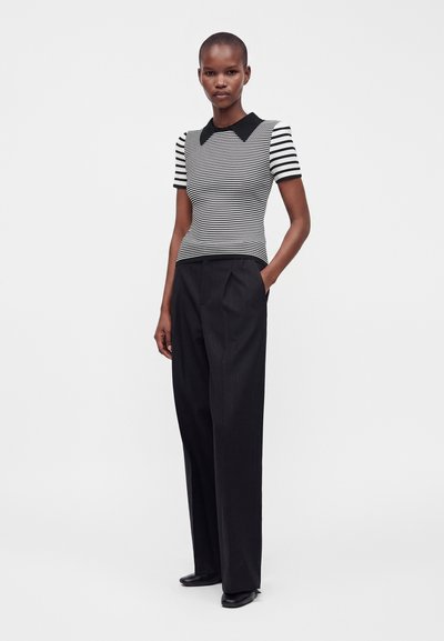 Femme portant un top à manches courtes rayé noir et blanc avec un col noir, un pantalon noir taille haute à jambes larges et des chaussures noires, debout avec une main dans la poche.