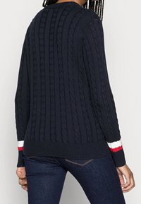 Pull en maille bleu marine avec motif torsadé texturé, encolure ronde et poignets côtelés ornés de rayures rouges et blanches. Coupe ajustée.