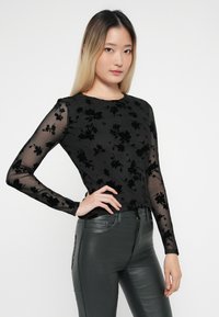 Sort langærmet top i gennemsigtigt stof med blomstret velourmønster, parret med mørke, skinnende bukser. Glat tekstur og tætsiddende design.