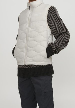 Person iført en hvid quiltet vest over en sort og grå sweater med chevronmønster og mørke cargobukser med hænderne i vestlommerne.