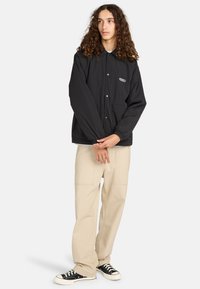 Zwarte lichtgewicht jas met drukknoopsluiting, elastische manchetten en zijzakken; gecombineerd met beige wijde broek en zwarte sneakers.