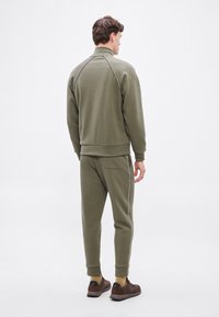 Σετ φόρμας σε ελιά πράσινο χρώμα, αποτελούμενο από δύο κομμάτια με ψηλό γιακά, διακοσμημένο με λευκές ρίγες, με rib τελειώματα στα μανίκια και τσεπάκι στο πίσω μέρος του παντελονιού.