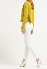 Un haut jaune à manches longues à nervures associé à un jean skinny blanc et des chaussures à talons hauts blanches. La vue de côté met en avant la silouhette détendue et l'ajustement.