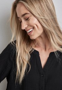 Camicetta nera con colletto a v, realizzata in tessuto leggero. Il modello ha lunghi capelli biondi lisci e un sorriso sottile, in profilo laterale.