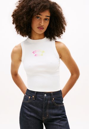 Giovane donna con capelli ricci che indossa un top corto senza maniche bianco con testo colorato "Tommy Jeans" e jeans di denim scuro a vita alta.