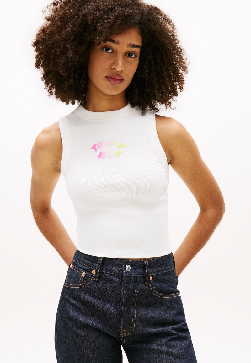 Νέα γυναίκα με σγουρά μαλλιά, που φοράει ένα αμάνικο λευκό crop top με πολύχρωμο κείμενο "Tommy Jeans" και ψηλόμεσα σκούρα τζιν παντελόνια.