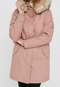 Parka rose clair avec capuche en fausse fourrure, équipée d'une fermeture éclair et de boutons-pression, deux poches avant, et une taille avec cordon de serrage pour un ajustement personnalisé.