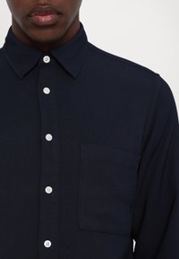 Homme portant une chemise bleu marine foncé à boutons blancs et une poche poitrine, visible des épaules jusqu'au milieu du torse.