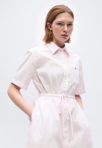 Νεαρή γυναίκα με κοντά ανοιχτό καστανά μαλλιά φοράει ροζ ανοιχτό ροζ φόρεμα με μανίκια κοντά, κουμπιά, ζώνη με κορδόνι στη μέση και τα χέρια στις τσέπες.