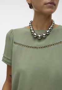 Blusa verde chiaro a maniche corte con dettagli in crochet sul colletto, abbinata a una collana di palline d'argento spessa.