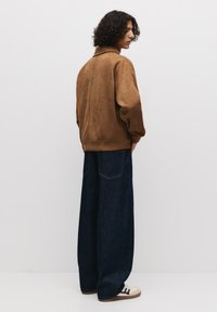 Manteau bomber en daim marron avec un ourlet côtelé, associé à un jean en denim foncé ample et des baskets blanches avec des rayures noires.