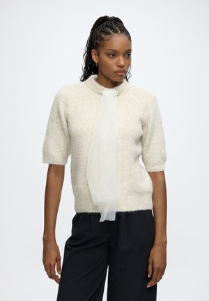 Femme aux cheveux tressés porte un pull crème à manches courtes avec une écharpe blanche transparente et un pantalon noir, debout devant un fond clair uni.