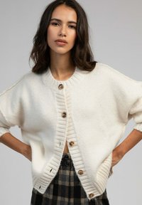 Cardigan en tricot couleur crème avec un col à côtes et de gros boutons, doté de manches amples et d'une coupe légèrement surdimensionnée.