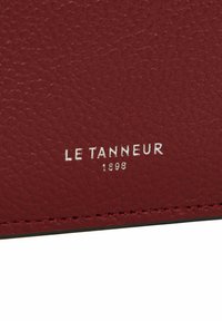 Prodotto in pelle bordeaux con superficie testurizzata e bordi cuciti, impreziosito da "LE TANNEUR 1898" in lettere d'argento.