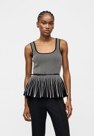 Femeie cu părul împletit, purtând un top fără mâneci în dungi alb-negru, cu tiv peplum și curea subțire neagră, asortat cu pantaloni negri.