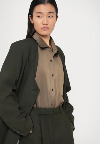 Blazer ajustat de culoare verde închis, cu umeri structurați, purtat peste o cămașă button-up de culoare bej deschis. Textură netedă și pantaloni ajustați completează lookul.
