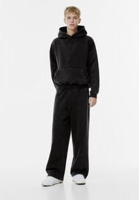 Čierna bavlnená mikina s kangaroo vreckom, voľný strih a rebrované manžety. Spárované s čiernymi teplákmi, oboje s elastickým pásom.