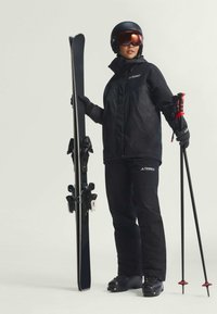 Čierna lyžiarska bunda a nohavice s logom, lyžiarske palice s červenými akcentmi, čierne lyže, prilba a okuliare. Hladký povrch a atletický strih.