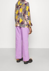 Blouse à motifs multicolores présentant des nuances de jaune, de violet et de noir, associée à un pantalon lavender ample et des sandales noires.