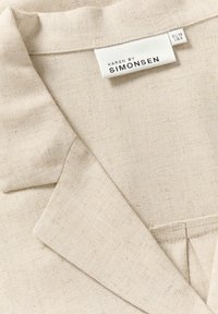 Béžový ľanový blazer s výstrihom do V, čistým šitím a štítkom s nápisom "KAREN BY SIMONSEN." Hladká textúra s jemnými škvrnami.
