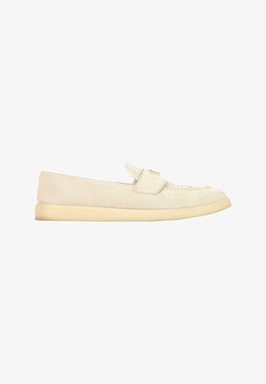 Beige suède loafer met een platte zool en een banden detail over de bovenkant, zijaanzicht op een witte achtergrond.